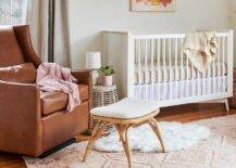 Vintage nursery design features a brown leather wingback glider with rattan stool and a white nursery crib with white and tan bedding atop a pink and gray nursery rug.