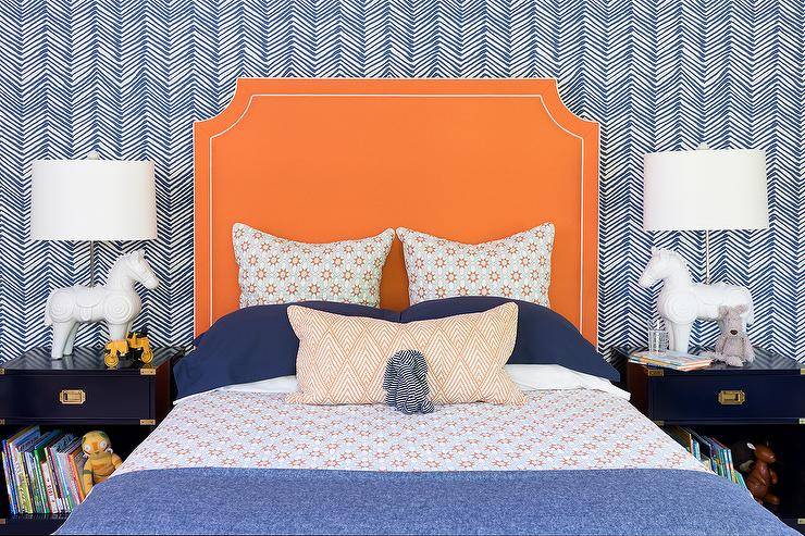 Orange and blue boy's bedroom features a wall covered in blue chevron wallpaper positioned behind an orange headboard lined with white piping. The bed is dressed in orange and blue bedding and flanked by blue campaign nightstands illuminated by white horse lamps.