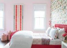 Red Jenny Lind spindle bed dressed with tassel shams flanked by white lacquer bedside tables in a girls bedroom. Colorful accent wallpaper gives a striking and bold appearance with a soft pink adjacent wall balancing the color and pattern.