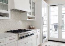 Kitchen features white cabinets with white and tan diamond pattern backsplash tiles and a white range hood.