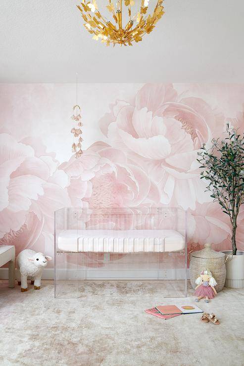 Nursery features a clear crib on a pink floral wall and a tall potted plant.
