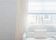 Light blue grasslcoth wallpaper frames a bedroom window dressed in ivory curtains hung from an acrylic and brass rod over a white and silver block print bench.
