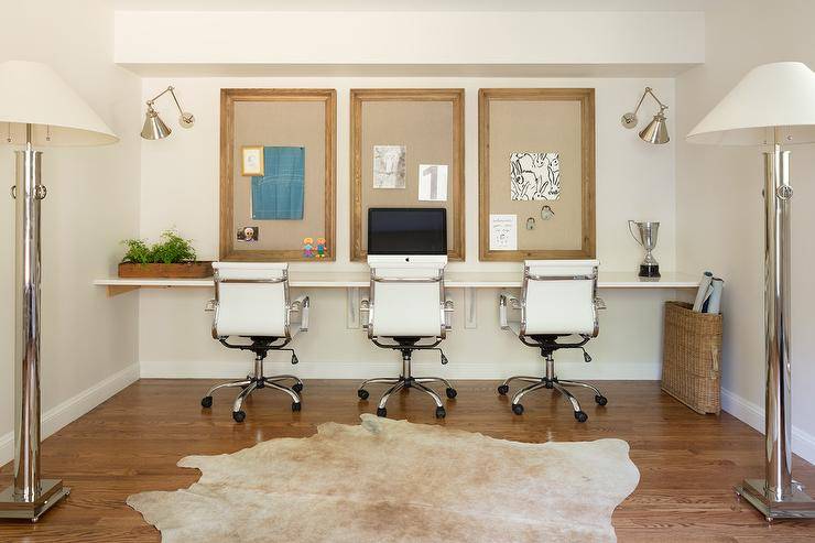 Homework room with a wall to wall desk features three task chairs, a framed pin board and sconces illuminating the space. A light brown cowhide rug layers over hardwood floors for a contemporary touch.
