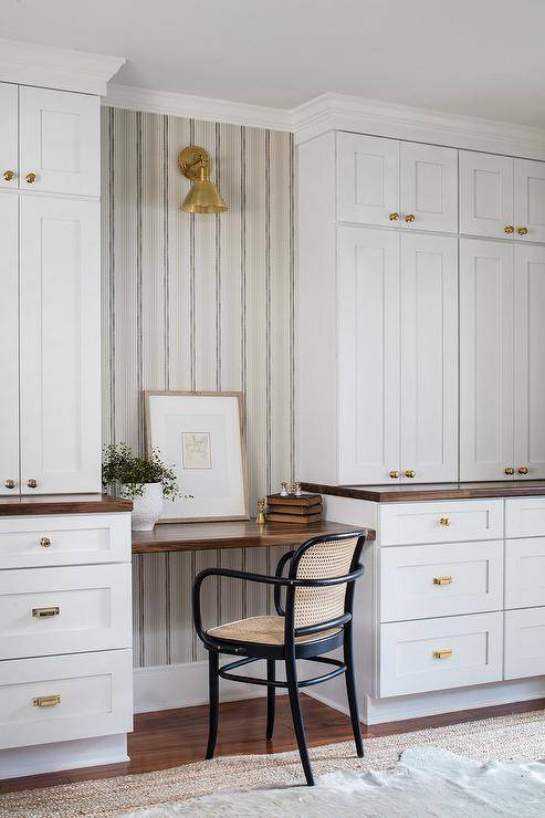 A tan and black cane chair sits at a wood floating desk on striped wall paper lit by a brass sconce flanked by white built in cabinets with brass knobs.