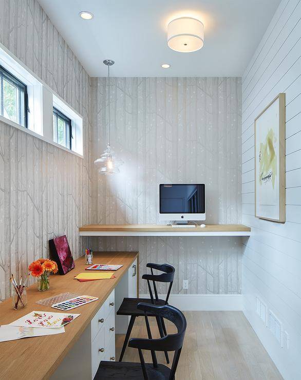 Long and narrow beautifully designed home office boasts walls clad in Cole & Son Woods Wallpaper lit by a drum flush mount. A wood top floating standing desk is mounted on an adjacent wall above a built in two-person two-tone desk accented with brass hardware and seating two black desk chairs. A window is located above the desk, while behind it, art hangs on a white shiplap accent wall.