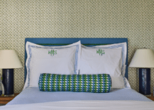 Green and blue monogrammed shams sit in front of a blue nailhead headboard and behind a blue and green bolster pillow placed on blue bedding. Blue lamps sit atop brown wooden nightstands.