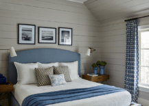 Blue and gray bedroom design features a blue upholstered bed with blue and white bedding flanked by X base bedside tables with blue flower vases lit by brass sconces on gray shiplap trim and a blue stripe rug with blue curtains.